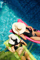 Wall Mural - Fit young women covering faces with straw hats when sunbathing on floating mattresses in hotel pool