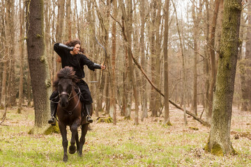 Wall Mural - beautiful black dressed woman in historical costume with bow and arrow shoots from a bow before riding a horse