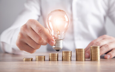 Wall Mural - Man holding light bulb. Stack of coins on the desk. Saving energy and money