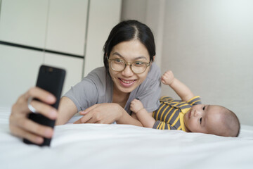 Wall Mural - Happy Smiling Asian Young mother and newborn baby boy lying on bed taking selfie with mobile phone together at home.