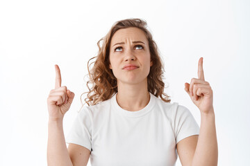 Skeptical and doubtful young blond woman having doubts, look with uncertain face at copyspace, pointing fingers up, standing over white background