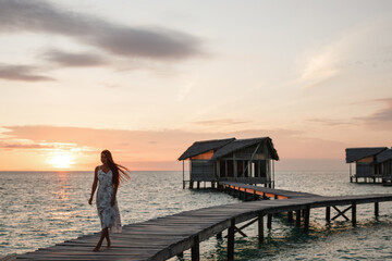 Wall Mural - Beautiful tourist woman in white dress on summer vacations walks over a wooden pier into the tropical sunset on the Maldives islands