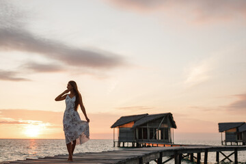 Wall Mural - A happy traveler woman walks on a wooden pier towards the beautiful sunset between water lodges on the Maldives islands