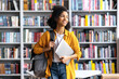 © Kateryna - Portrait of joyful pretty african american female student standing against background of bookshelves in university library holding laptop and backpack looking to the side, smiling pleasant
