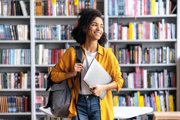 portrait of joyful pretty african american female student standing against background of bookshelves