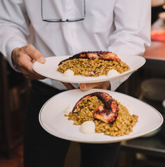 Camarero de un restaurante de lujo llevando dos platos gourmet de arroz con tentaculo de pulpo