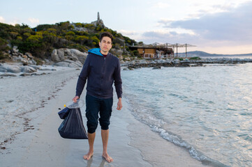 Portrait of young volunteer man looking at the camera while standing on the beach shore with barefoot holding a bag full of waste.