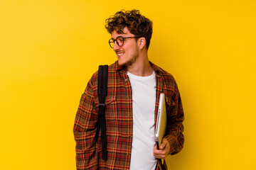 Young student caucasian man holding a laptop isolated on yellow background looks aside smiling, cheerful and pleasant.