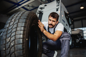 Wall Mural - Young dedicated hardworking mechanic crouching in garage of import and export firm and preparing to change tire on truck.