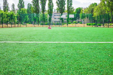 little girl sitting in the middle of the soccer field with artificial green grass near the school. a