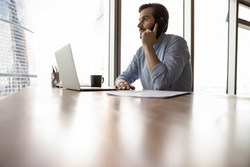 Wall Mural - Successful young Caucasian businessman sit at desk in modern office building work on laptop talk on smartphone with business partner. Confident man have cellphone call with client or customer.