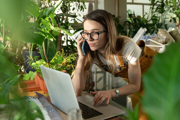 Wall Mural - Freelance female work from home, woman gardener use laptop while talking with client on smartphone. Small floral business owner, florist entrepreneur make order online in home office at indoor garden