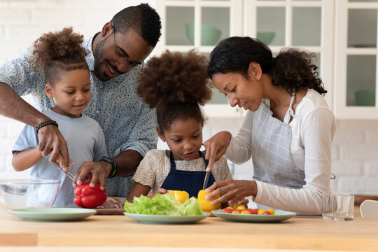 happy family couple teaching two preschooler kids preparing salad. parents and children cooking dinn