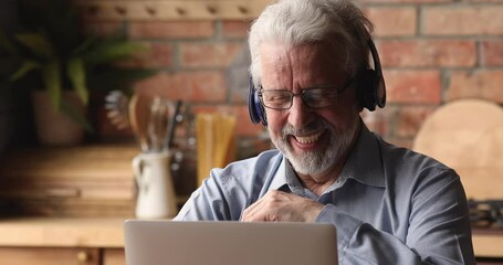 Wall Mural - Head shot happy middle aged senior hoary man in eyeglasses wearing wireless headphones, enjoying videocall zoom web camera internet meeting using computer, communicating distantly online in kitchen.