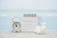White Calendar And Alarm Clock On Concrete Floor With Blurred Plumeria Flower And Seascape Background For Recreation Or Work From Home Concept