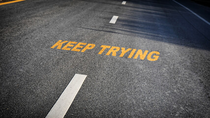 Keep trying word with white dividing lines on black asphalt road surface with sunlight, business challenge concept and effort idea