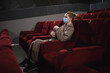 © Aleksandra Iarosh - young woman in medical mask eating popcorn sitting in the cinema