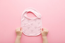 Baby Girl Hands Holding Textile Bib With Heart Shapes For Feeding On Light Pink Table Background. Pastel Color. Closeup. Point Of View Shot. Top Down View.