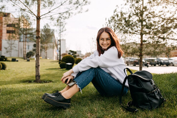 Wall Mural - Young happy female student sitting on grass in park, with a backpack, smiling.