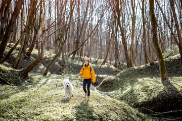 Wall Mural - Young woman in hiking clothes and backpack spend time together with big white dog in green spring forest. Enjoys and explore of tranquil nature.