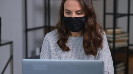 Wall Mural - Pensive young businesswoman wearing protective medical mask sitting on the workplace in front of the laptop, looks away and worrying. Female employee in medical mask