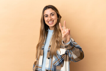 Wall Mural - Young caucasian woman isolated on beige background smiling and showing victory sign