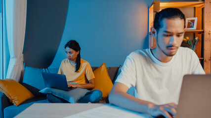 Canvas Print - Freelance asia couple man and woman in casual feel serious focus on work separate laptop computer at night, hasband sit in front on table wife sit behind on couch at house. Work from home concept.