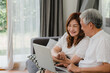 © tirachard - Asian senior couple video call at home. Asian Senior Chinese grandparents, using laptop video call talking with family grandchild kids while lying on sofa in living room at home concept.