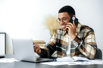 Wall Mural - Satisfied smiling mixed race latino male freelancer or manager with glasses, have cell phone conversation, sits at work desk, uses laptop, checking e-mail, looking for information at internet