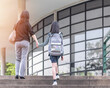 © Chinnapong - Educational back to school, parent's day, bring kid to work concept with elementary student girl child carrying backpack holding mother's hand walking up admission office building going to class