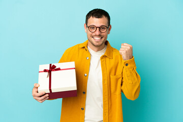 Poster - brazilian man holding a gift over isolated blue background celebrating a victory in winner position