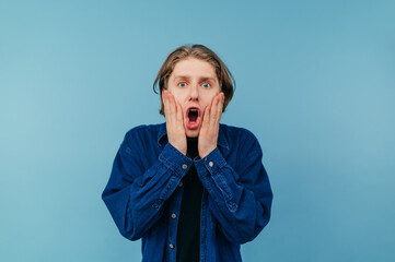 Wall Mural - Surprised guy in casual clothes stands on a blue background and putting his hands to his face emotionally looks at the camera and screams in shock.