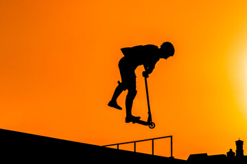 Wall Mural - Unrecognizable teenage boy silhouette showing high jump tricks on scooter against orange sunset sky at skatepark. Sport, extreme, youth, urban culture, freestyle, outdoor activity concept