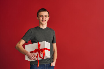 young guy, a Caucasian teenager in casual clothes holds a large white gift box with a red ribbon and smiles on a red background. The concept of giving gifts, winning