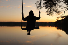 Silhouette Of A Romantic Young Woman On A Swing Over Lake At Sunset. Young Girl Traveler Sitting On The Swing In Beautiful Nature, View On The Lake