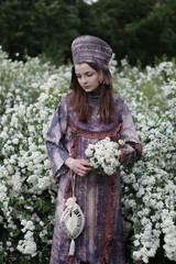 Wall Mural - Look of stylish russian girl in traditional national folk dress (sundress) and slavic headdress (kokoshnik) in garden with spring white flowers. Russian fashion. Spring nature, seasonal bloom, blossom