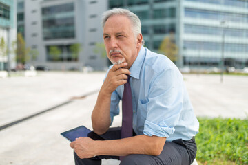 Canvas Print - Businessman using his tablet outdoor while sitting on a bench in front of his office