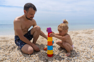 Young caucasian man with little child play with colorful toys at sandy beach near the sea, summer time, tropical resort, father and daughter make pyramid, happy fathers day.