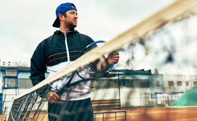 View through the net of a male tennis player playing on a tennis court. Young man tennis player playing match outdoors.