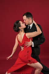 Couple of professional tango dancers in elegant suit and dress pose in a dancing movement on red background. Handsome man and woman dance looking  eye to eye.