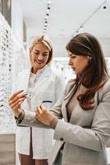 Sticker - Beautiful and fashionable woman choosing eyeglasses frame in modern optical store. Female seller specialist helps her to make right decision.