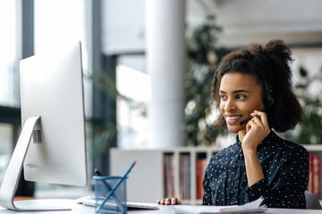 Confident beautiful african american girl in headset, manager, call center worker or freelancer, sitting at the table, using computer, communicating with colleague or client via video link, smiling