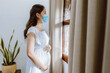 © Oulaphone - Young pregnant Asian woman wearing medical mask standing near window at home for self quarantine from spread of Covid 19 diseases.