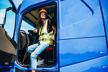 Portrait of beautiful young woman professional truck driver with protective yellow helmet sitting and driving a big truck. Inside of vehicle. People and industrial transportation concept.