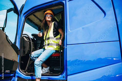 Portrait of beautiful young woman professional truck driver with protective yellow helmet sitting and driving a big truck. Inside of vehicle. People and industrial transportation concept.