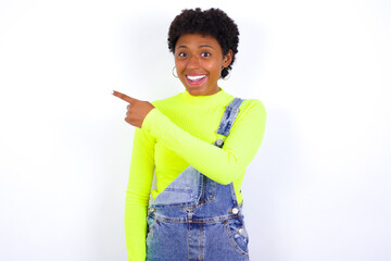 Wall Mural - young African American woman with short hair wearing denim overall against white wall pointing away and smiling to you. Look over there!