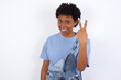 © Roquillo - African American female with afro hair wears jeans overalls over white wall smiling and looking friendly, showing number two or second with hand forward, counting down
