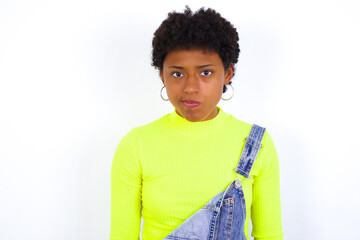 African American female with curly bushy wears jeans overalls over white wall Pointing down with fingers showing advertisement, surprised face and open mouth