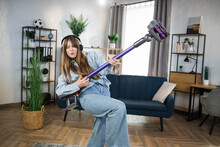 Positive young woman listening music in wireless headphones and playing with handheld vacuum cleaner. Housekeeping with fun. Modern devices concept.