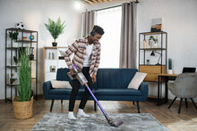 Smiling Afro American Man In Wireless Headphones Cleaning Carpet With Handheld Vacuum Cleaner. Young Guy Enjoying Housekeeping Using Modern Devices.
